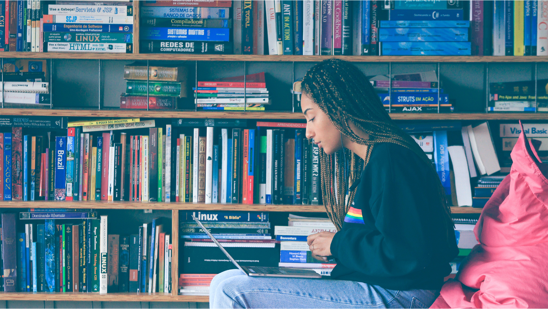 Uma mulher com tranças, sentada, usando um laptop em frente a uma estante cheia de livros.