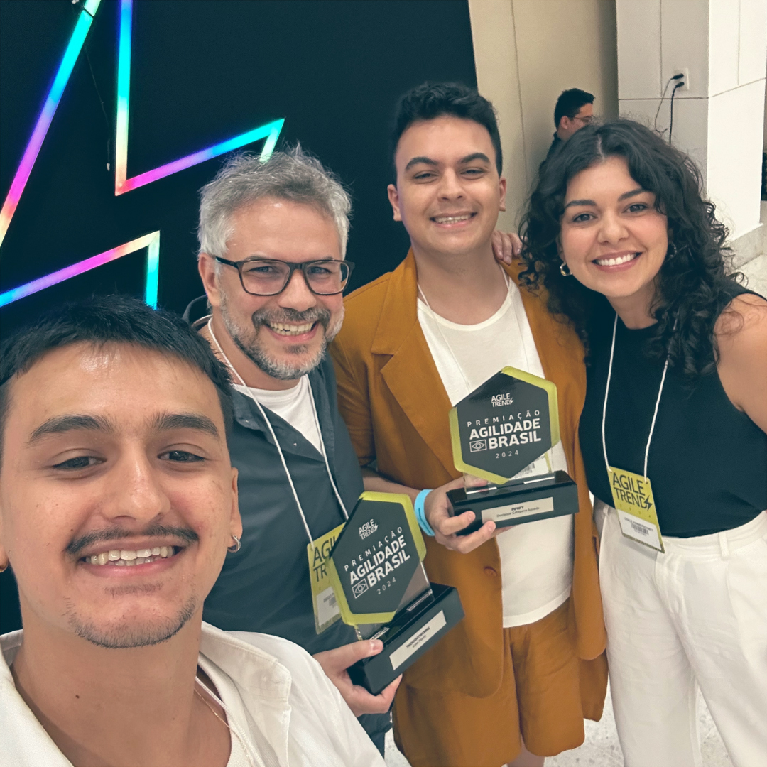 Un equipo celebrando en el evento Prêmio Agilidade Brasil 2024, sonriendo mientras sostienen sus premios. Llevan credenciales del evento y están de pie frente a un fondo iluminado con neón.