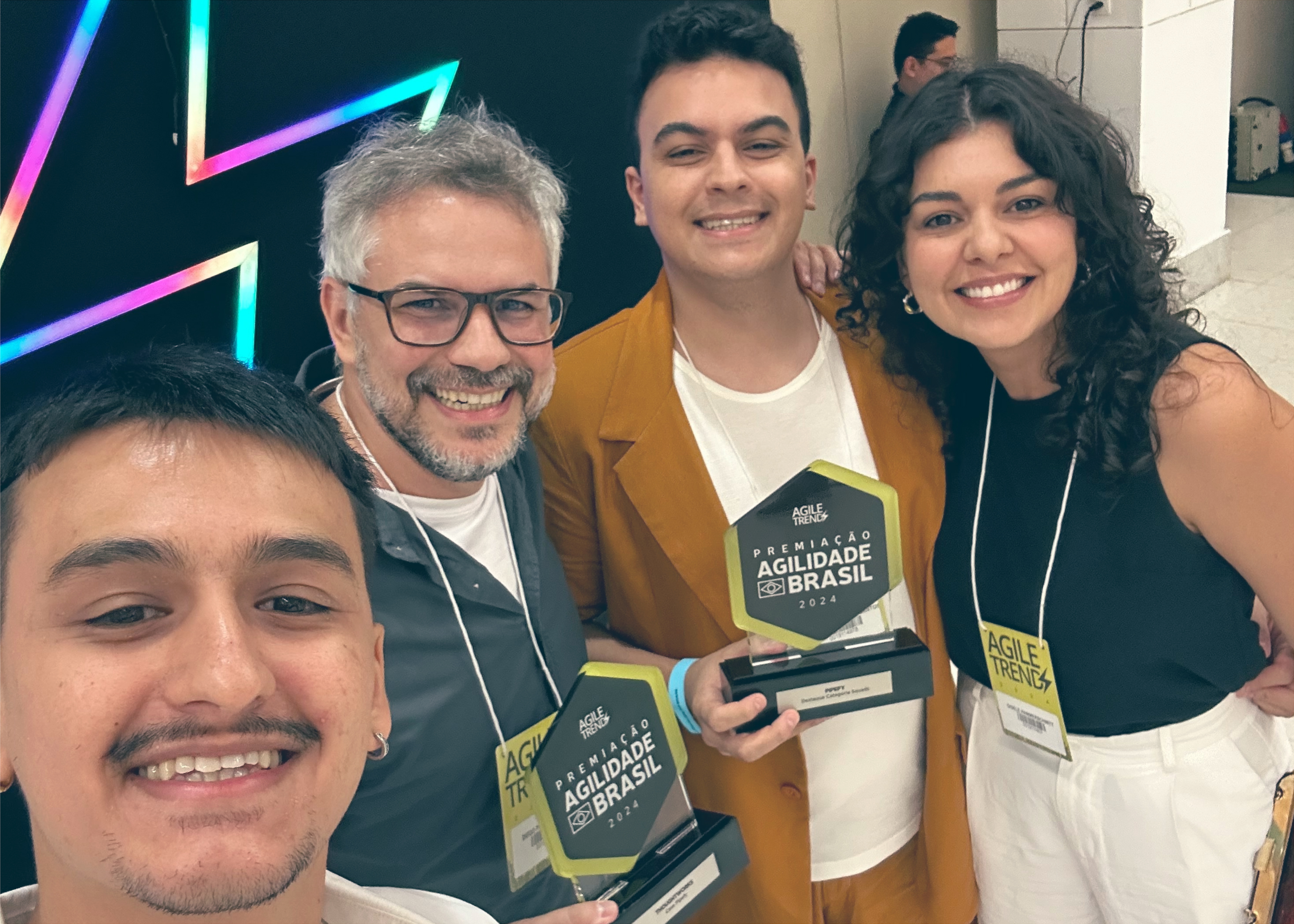 Un equipo celebrando en el evento Prêmio Agilidade Brasil 2024, sonriendo mientras sostienen sus premios. Llevan credenciales del evento y están de pie frente a un fondo iluminado con neón.